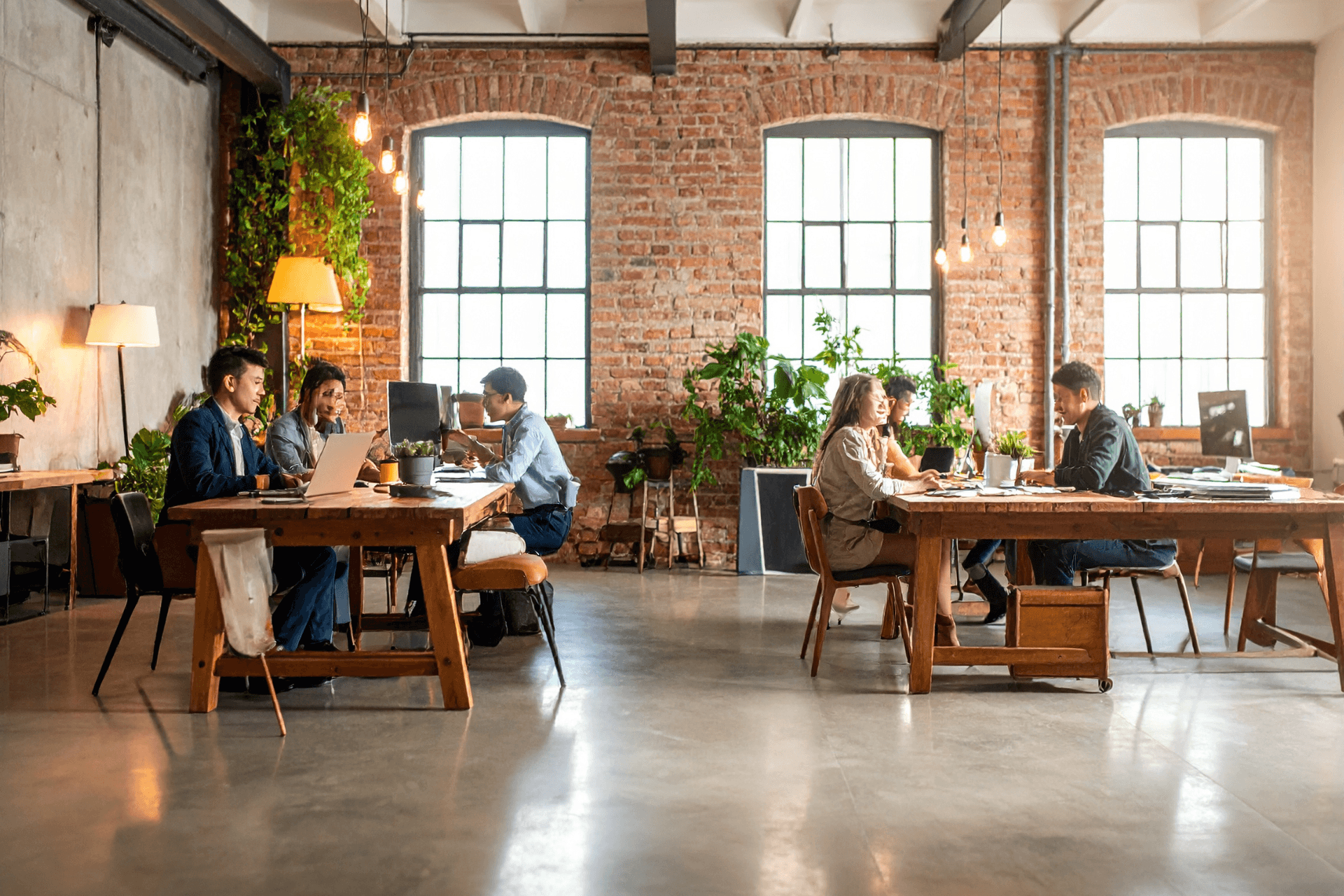 Teams arbeiten an langen Holztischen in einem Loft mit Ziegelwänden und hohen Fenstern